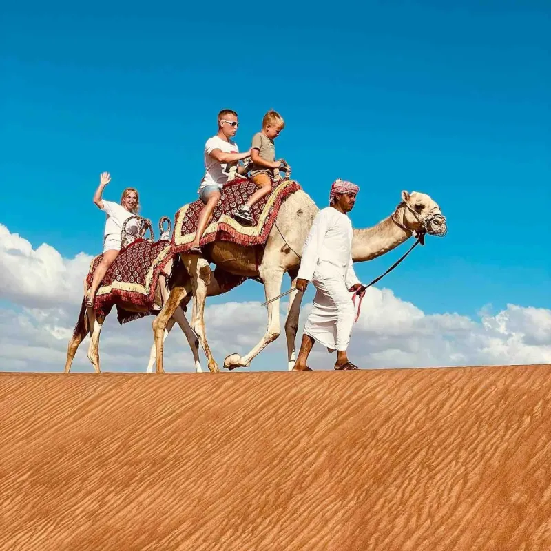 Family riding camels led by a guide across the red dunes